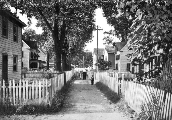 View of four men and a boy walking down a narrow lane lined with homes and a picket fence.