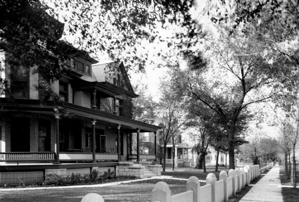 View of a row of homes in the residential district. The brick house in the foreground features a large wraparound porch and a fence around the front yard.