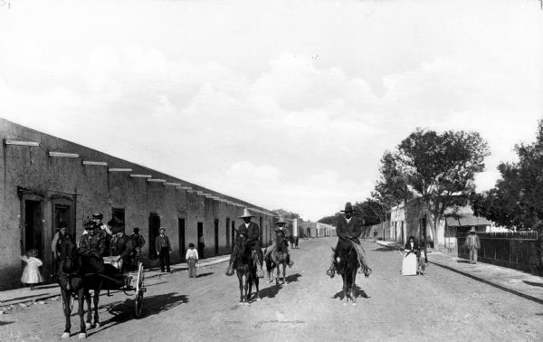 People ride on horseback and in carriages down a dirt road.  A long adobe building can be seen on the left.