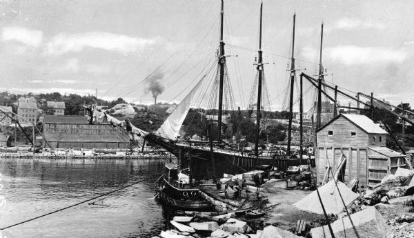 View of the docks at Rockport Granite Company, operating from 1864-1927.  A sailing ship can be seen in the water near industrial buildings.