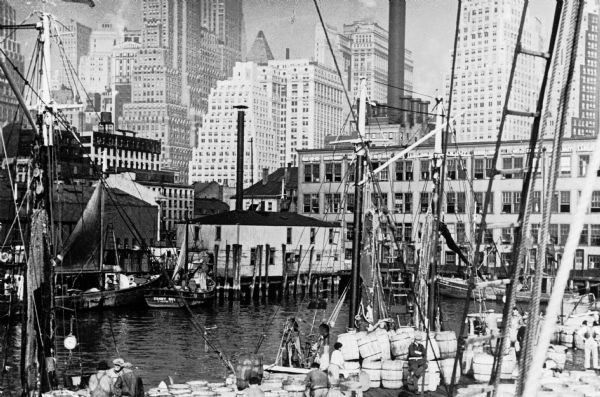View of the wharf, an area crowded with boathouses, fishermen, and barrels of goods. The city's skyscrapers rise in the background.