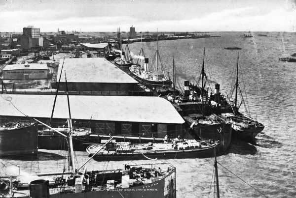 View of Galveston Harbor.  Ships are stationed near large piers and warehouses.  Published by Chas Daferner.