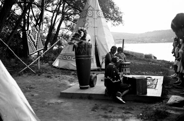 An American Indian family demonstrates their craft before a group of young girls. A tipi can be seen along the shore of the Hudson River, and a traditional blanket hangs on a clothesline.