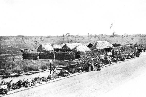 View of a Seminole Indian village.  A family walks past huts, surrounded by fences.