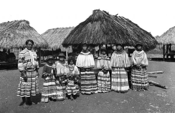 Portrait of a group of Seminole Indians.  Children and adults stand before huts in their village, wearing  traditional clothing.