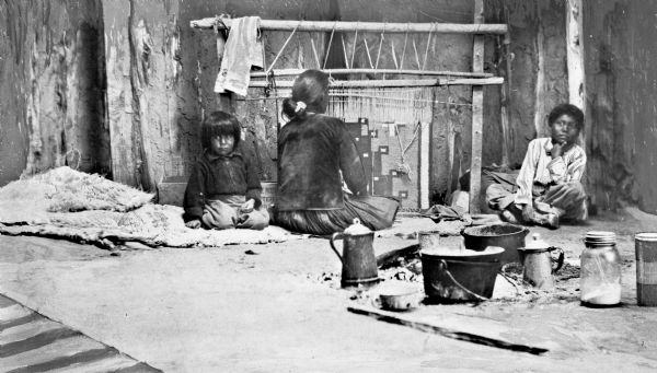 A seated Navajo woman weaves a blanket near two young children. Cooking pots and utensils are in the right foreground.