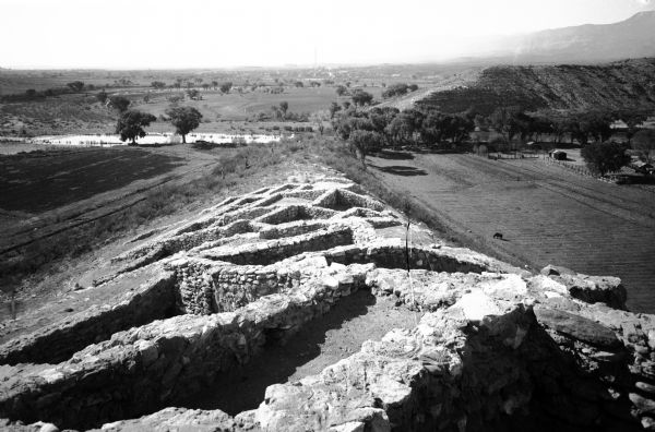 View of the land around Pueblo living quarters; the quarters are a stone and mortar construction that rises to a central rectangular building.