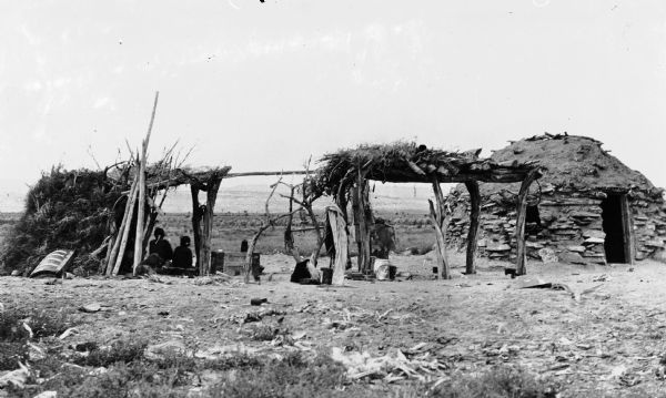 Two women sit near Navaho hogans and a brush arbor.