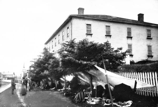 View of a market near a road.  Native Americans sell baskets and curios to men and women wearing hats and long coats.