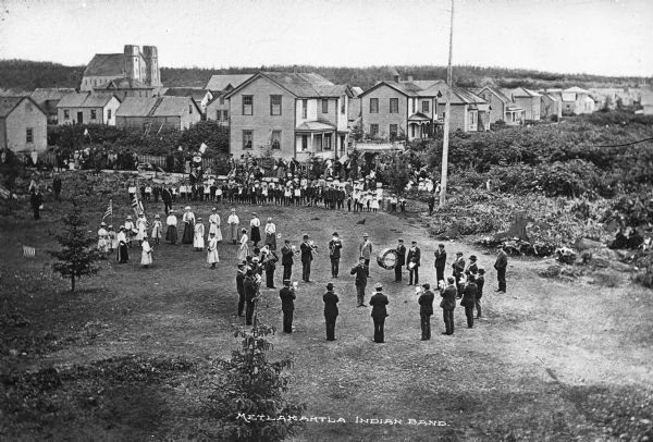 View of the Metlakahtla Brass Band as they perform in a circular formation around the conductor. Another group of women and girls stands to the left. Spectators stand near rows of houses.