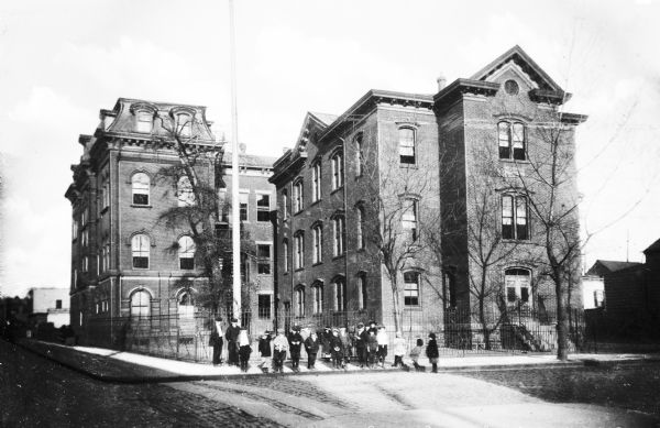 Children stand on the sidewalk outside Public School Number Six, a tall brick building on a city block.