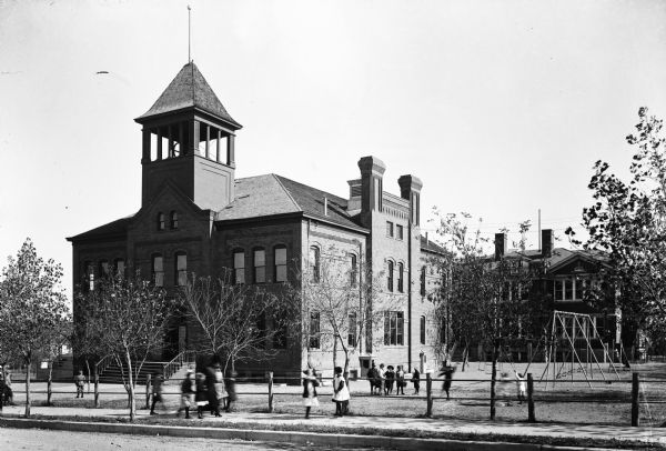 Exterior of Central Ward School, a brick building featuring a central colonnaded tower.  Children can be seen near the playground outside the school.
