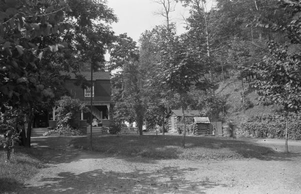 Exterior of a country home in the woods. Men and women sit on the porch of the house, and a wooded hill rises on the right.