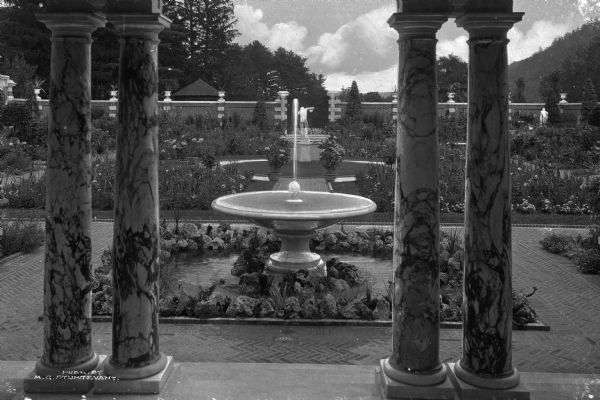 View of the Italian garden at Brookside, the luxurious estate of Mr. W.H. Walker, through the porch columns.  A fountain and sculpture can be seen amidst the flowers and other plants in the garden, designed by landscape architect Ferruccia Vitale.