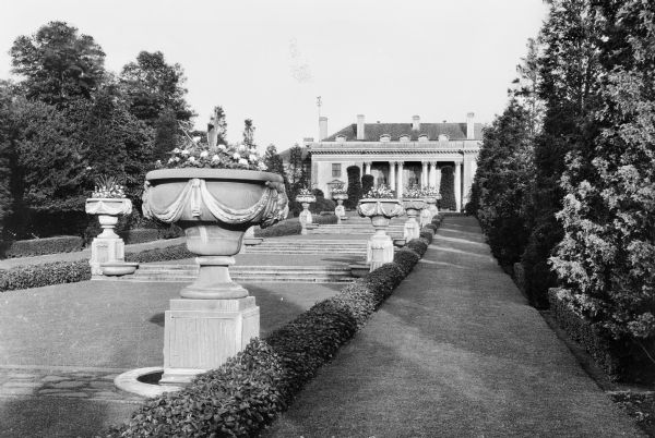 View of Nemours, one of the palatial estates of the Alfred I. du Pont (1864-1935) family built between 1909-1910 by Smyth and Son. Side lawn paths frame a center lawn lined with stone flower urns on both sides. A series of stone steps, broken up by the lawn, lead up to the mansion.