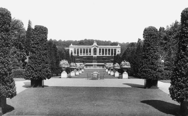 View of Nemours, one of the palatial estates of the Alfred I. du Pont (1864-1935) family built between 1909-1910 by Smyth and Son. A staircase leads through a garden, lined with flower vases.  A colonnaded facade stands in the distance.