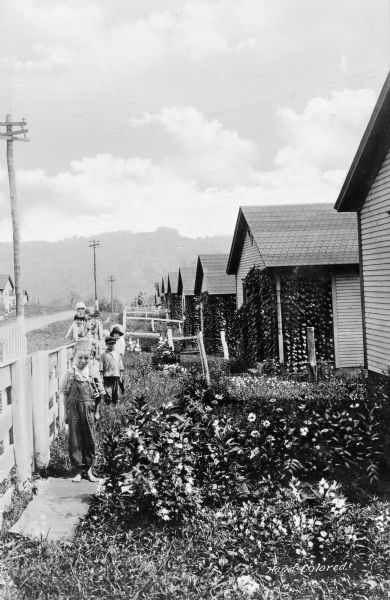 A group of children stand along a fence near a row of cottages at the outskirts of town. In the far background is a mountain range.