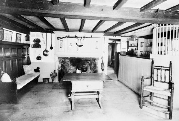 Interior view of Longfellow's Wayside Inn, which opened in 1716. The Old Bar Room features a wooden bar counter, a fireplace with a hunting rifle above, and a beamed ceiling.