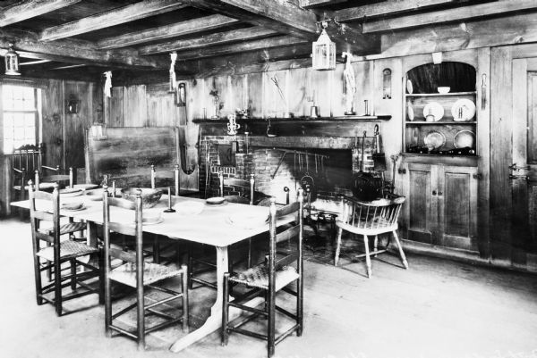 Interior view of Longfellow's Wayside Inn, which opened in 1716. The Old Kitchen features a large fireplace, wood paneled walls, beamed ceiling, and a long wooden table with chairs.