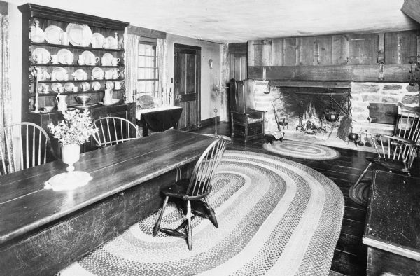 Interior of the living room of an Early American home, built 1765.  A long wooden dining table stands near a wooden hutch and a fireplace.  Large braided rugs can be seen on the floor.