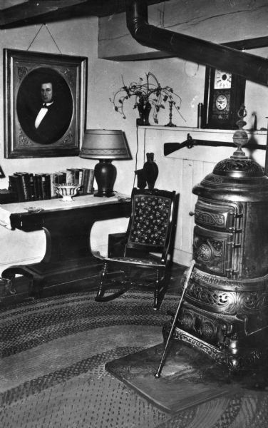 Interior view of a farmhouse.  The living room features a potbelly stove and a rocking chair.  The portrait of a man hangs on the wall above a table lined with books.