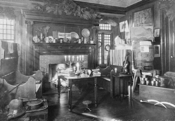 Interior view of an early American home.  The room features an elaborate fireplace, a violin, and paintings.