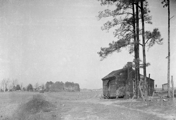 Exterior view of a small cabin located near a cotton field. There is a cemetery in the far background.