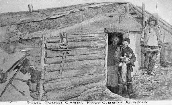 Exterior of a cabin with its occupants posed in the doorway. One of the men is holding a dog and smoking a pipe.
