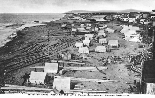 Elevated view of an Eskimo tent village on sandspit along the shoreline. Wood buildings are along higher ground on the right near a river.
