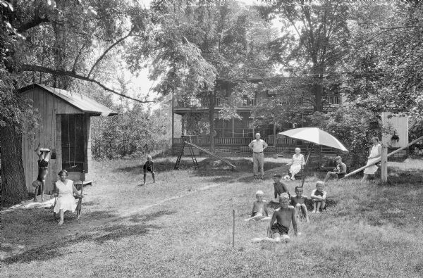 Posed portrait of a large family in the yard in front of their home. Four adults pose with children who are playing on swings, sitting in the shade under an umbrella, and posing with dogs. There is a slide made out of a ladder, and a teeter-totter.
