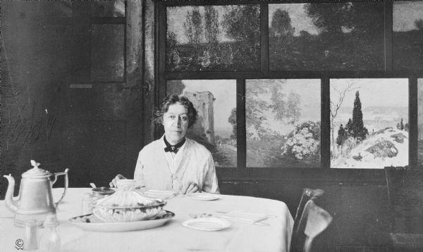 View of a woman, Miss Florence, seated at a dining room table.  Landscape paintings are on the paneled wall behind her.