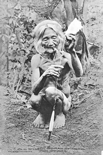 Portrait of an elderly Ifugao woman squatting on the ground using a drop spindle for spinning in the Philippines. She is holding something in her mouth.