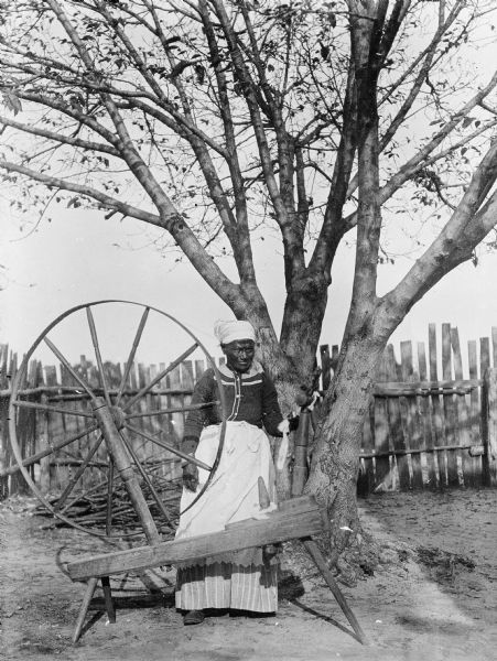 View of a woman standing near a tree in the yard with a large spinning wheel.