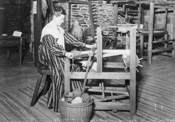 View of a woman weaving with a loom as an exhibit at the Farmer's Museum, which opened in 1944. There is another loom behind her.