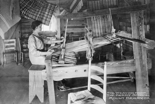 View of a woman weaving at a loom in the weaving room of the Berry School, which opened in 1902. Blankets are hanging near the ceiling.