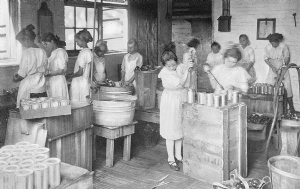 Interior of a company school operated by Gainesville Cotton Mills.  Girls in the room work over large tubs and crates with cans and canning equipment. Boxes of fruit or vegetables sit on the floor.