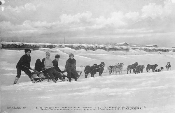 View of four men of the U.S. Signal Crew out on a dogsled to repair a break in a telegraph line. There are cabins in the background. Copyright by A.J. Johnson.