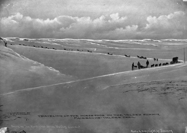 View from a distance of a pack train winding up a mountain on the Fairbanks-Valdez Trail. A man pushing a bicycle is at the end of the group. Copyright by A.J. Johnson.