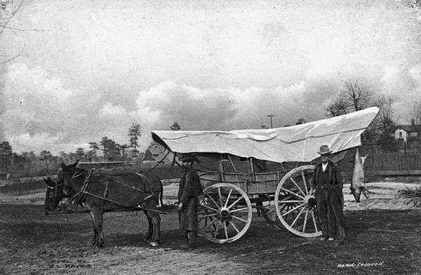 View of two men standing near a Sand Hill Supply Company mule-drawn Conestoga-type wagon with a slaughtered pig hanging on the back. Published by C.L. Hayes.