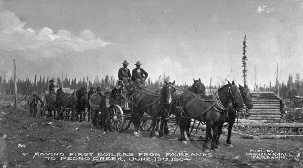 View of men moving boilers on horse-drawn wagons from Fairbanks to Pedro Creek, June 13th, 1904. Caption reads: "Moving First Boilers From Fairbanks To Pedro Creek, June 13th, 1904." and "Publ. By Chisholm & Hall, Fairbanks."
