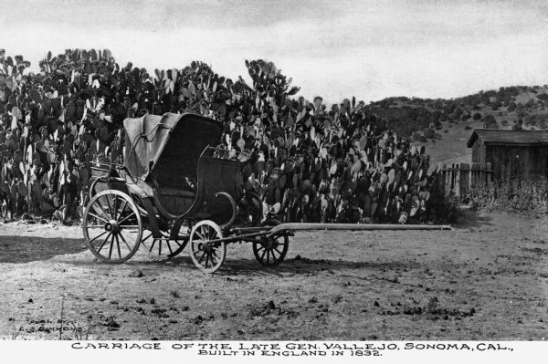 View of the carriage of the late General Vallejo, which was built in England in 1832. A field of cacti can be seen behind the carriage. There is a fence and outbuilding on the right, and a mountain range in the background. Published by L.S. Simmons.