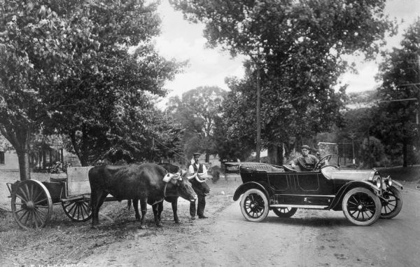 View of an man standing with an ox-drawn cart near a man sitting in an automobile.