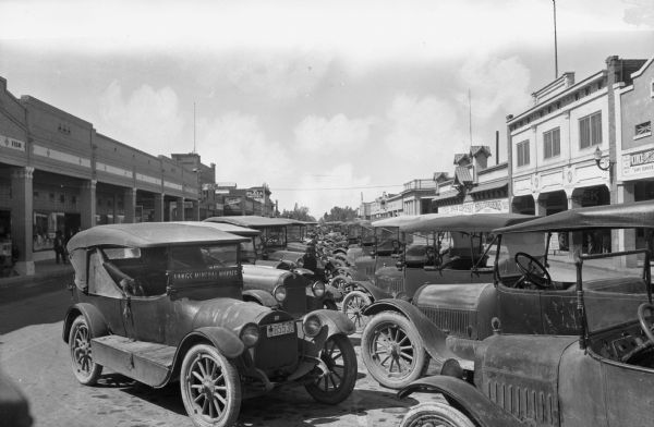View down rows of automobiles lined up and parked at an angle in the middle of the street. Sidewalks and storefronts are on both sides of the street.
