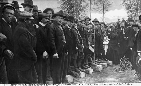 A crowd gathers as a golden stake is driven as a part of a ceremony to celebrate the completion of the Tanana Valley Railroad. Caption reads: "Driving Golden Spike, Tanana Valley, RY, Fairbanks, Alaska."