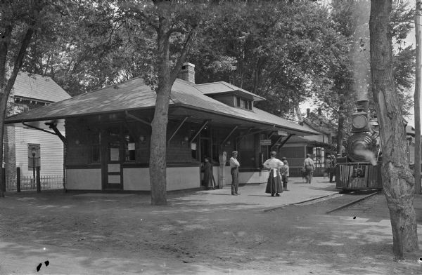 Exterior view of Barnegat Railroad Station. A locomotive is in front of the platform, and children and adults stand near the railroad tracks.
