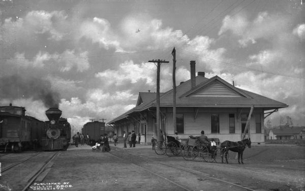 Exterior view of a railroad depot. Men and women gathered on and near the tracks to board trains. Published by Quillin Bros. Druggists.