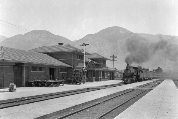 Exterior of Provo Railroad Station.  Baggage trucks can be seen near the tracks as a train arrives at the station.