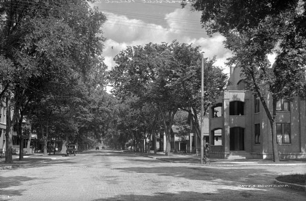View down Broad Street. There is one automobile parked on the left, and some horse-drawn vehicles in the far distance. Houses and sidewalks are on both sides of the street.