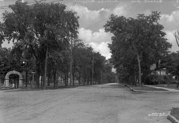View of entrance to White Park from the intersection of College Street and Bushnell Street, which are unpaved. A young girl stands under the archway entrance to the park. Across the street a dog is in the front yard of a house.