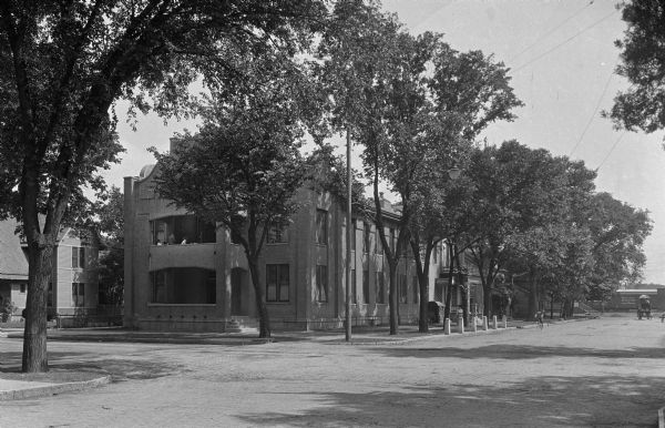 View of the Beloit Hospital from an intersection. There are people sitting on a second floor balcony in a building on the corner. A person is riding a bicycle down the street and a woman is walking down the sidewalk. At the end of the street are railroad cars, and a horse-drawn wagon.